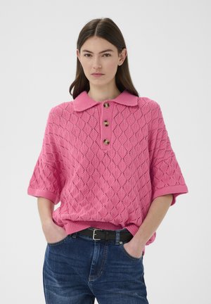 Vrouw draagt een roze gebreide korte mouwen trui met drie knopen en kraag, gecombineerd met een blauwe spijkerbroek en een zwarte riem, handen in de zakken.