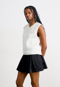 Maglione smanicato bianco a maglia cable knit con scollatura a V e ricamo blu, abbinato a una gonna nera plissettata, in piedi su uno sfondo neutro.