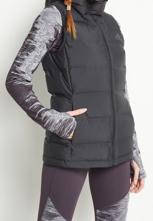 Femme portant une veste sans manches matelassée noire par-dessus un top gris à manches longues à motifs et des leggings assortis, main dans la poche de la veste, debout sur fond blanc.