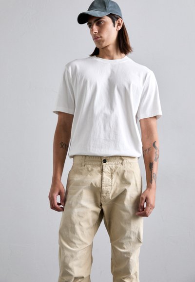 Tânăr îmbrăcat cu o șapcă gri, un tricou alb și o pereche de pantaloni bej, stând cu tatuaje vizibile pe ambele antebrațe, pe un fundal simplu deschis.