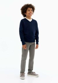 Maglione navy a collo a V in un tessuto morbido, abbinato a jeans grigio chiaro e scarpe da ginnastica grigie. Il bambino sorride con i capelli ricci.