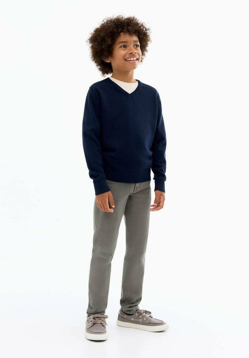 Maglione navy a collo a V in un tessuto morbido, abbinato a jeans grigio chiaro e scarpe da ginnastica grigie. Il bambino sorride con i capelli ricci.
