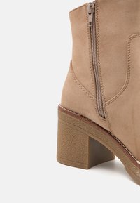 Beige ankelstövel i mocka med fyrkantig tå, sidosdragkedja och texturerad blockklack. Har synliga sömmar och en lätt kurva längs sidorna.