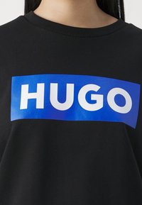 Zwarte sweatshirt met een blauw rechthoekig grafisch ontwerp en de witte tekst "HUGO" prominent op de voorkant weergegeven.