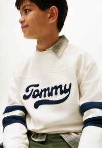 Crème sweatshirt met marineblauwe strepen, voorzien van een geborduurd "Tommy" logo. Gedragen over een geruite kraagshirt in grijstinten.