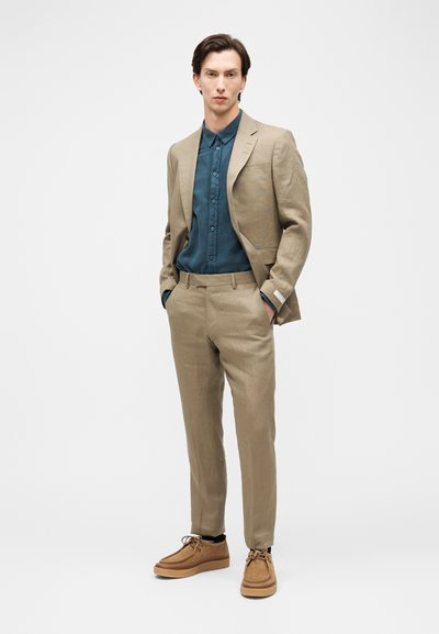 Νεαρός άνδρας όρθιος με τα χέρια στις τσέπες, φοράει ένα μπεζ κοστούμι, μπλε-πράσινο πουκάμισο με κουμπιά και καφέ παπούτσια από σουέτ με κορδόνια, σε απλό φόντο.