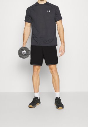 Mustat urheilushortsit ja tummanharmaa lyhythihainen paita, valkoiset sukat ja mustat lenkkarit, pidellen pyöreää mustaa käsipainoa.