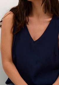 Mörkblå linne med V-ringning som är tillverkat av en mjuk textil. Har smala axelband och en avslappnad passform som faller något över axlarna.