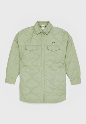 Chaqueta de camisa acolchada de color verde claro con un cuello puntiagudo, botones a presión, dos bolsillos en el pecho y pequeño logo negro de Nike en el lado izquierdo.