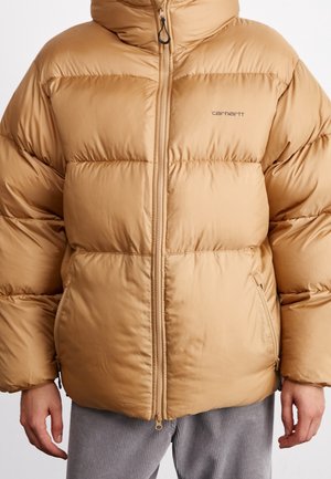Doudoune beige avec fermeture éclair frontale et poches zippées, modèle portant un pantalon gris, logo Carhartt sur le côté gauche de la poitrine.