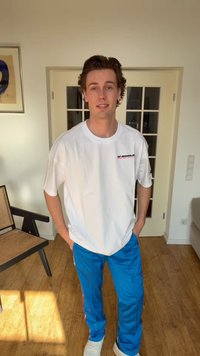 Mladý muž stojaci v interiéri, oblečený v bielom tričku Kappa, modrých nohaviciach a bielych teniskách, s rukami v kapsách v miestnosti s drevenou podlahou.