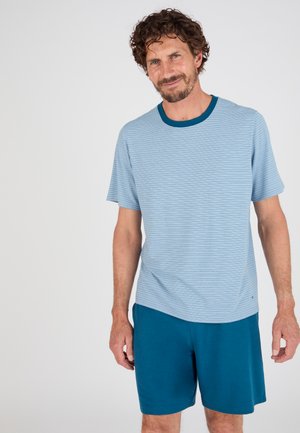 Mann mit lockigem Haar, der ein hellblau gestreiftes T-Shirt und dunkelgrüne Shorts trägt, steht vor einem schlichten weißen Hintergrund.