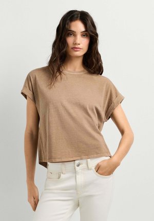 Vrouw met golvend donker haar, gekleed in een losse lichtbruine top met korte mouwen en witte broek, staand met één hand in de zak.