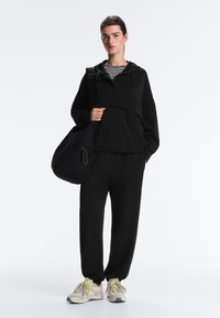 Osoba v čiernej kapucni a nohaviciach, pod ktorou má pruhované tričko, nesúca veľkú čiernu tašku cez jedno rameno, obutá v bielych teniskách so žltými šnúrkami.