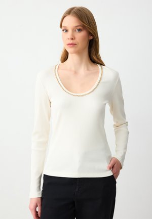 Top bianco a maniche lunghe con scollo ampio decorato da un bordo in catena dorata. Il tessuto ha una texture morbida, abbinato a pantaloni scuri.