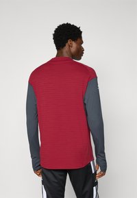 Chemise à manches longues bordeaux avec un motif texturé sur le haut, manches grises et col rond. Portée avec un pantalon noir à rayures blanches.