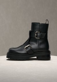 Bottine noire en cuir avec semelle épaisse et deux larges brides, chacune sécurisée par des boucles en métal argenté, sur un fond neutre.