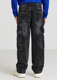 Pantaloni cargo in denim nero con una texture sbiadita, dotati di multiple tasche e una vestibilità ampia, indossati con una camicia a maniche lunghe blu e sneakers bianche.