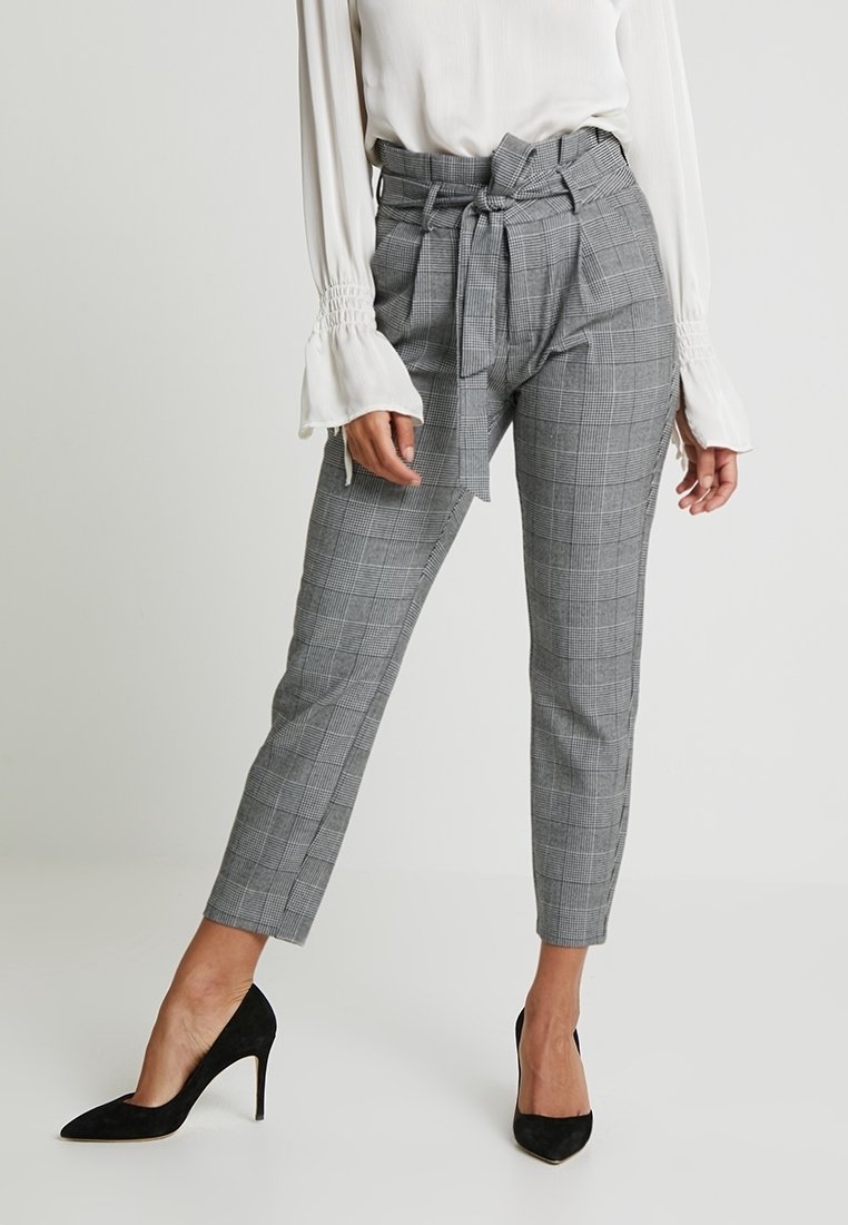 Femme portant un pantalon à carreaux gris taille haute avec une ceinture nouée, associé à un chemisier blanc à manches longues et des talons hauts noirs.