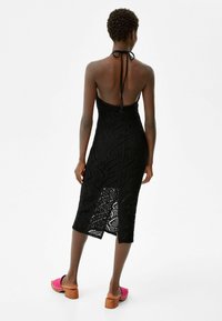 Robe en crochet noire avec un col halter et un dos nu, présentant une silhouette longueur genou et une texture ornée de motifs en feuilles.