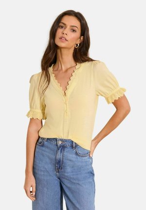 Woman wearing a pale yellow short-sleeve top with scalloped lace trim and blue high-waisted jeans, posing with one hand on hip.