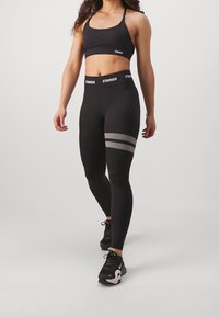 Crop top sportivo nero con spalline sottili, abbinato a leggings neri a vita alta con tre righe punteggiate orizzontali sulla coscia.