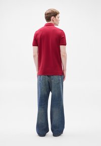 Κόκκινο πόλο πουκάμισο με κοντά μανίκια, συνδυασμένο με χαλαρά μπλε τζιν. Το πουκάμισο έχει γιακά και πλευρικές σχισμές στο τελείωμα.