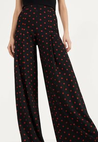 Löysät housut, jotka on valmistettu teksturoidusta mustasta kankaasta, jossa on punaisia pilkkuja. Housuissa on korkea vyötärö ja terävät poimut lisäämään muotoa.