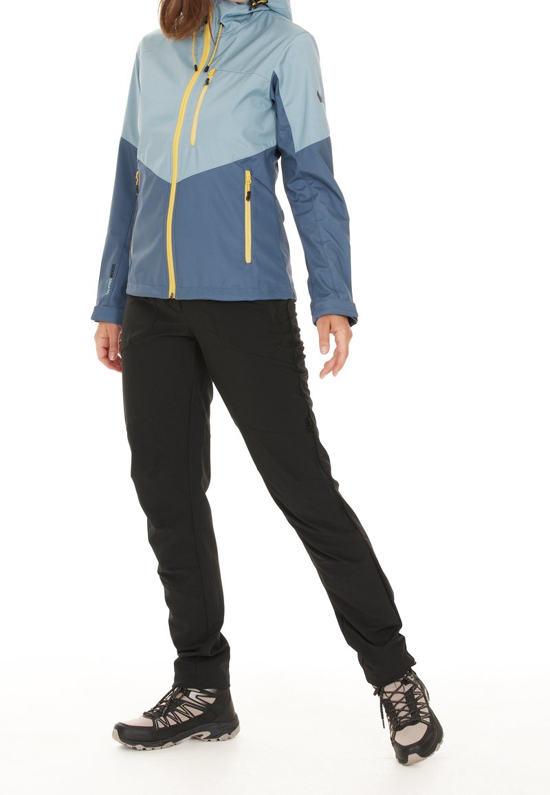 Personne portant une veste zippée bleue et marine avec des tirettes jaunes, un pantalon noir et des chaussures de randonnée noires, debout, jambes écartées.