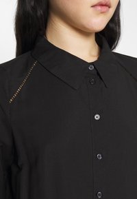 Femme portant une chemise noire à boutons avec un col pointu et des coutures décoratives le long des coutures des épaules.