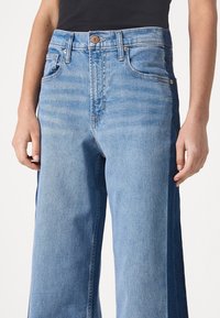 Ljusblå denimjeans med vida ben, med fem fickor och en knappknäppning. Distinkt färgvariation längs sidsömmarna.