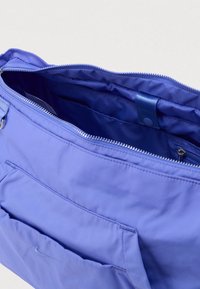 Ανοιχτή ανοιχτό μωβ τσαντάκι με φερμουάρ που αποκαλύπτει πολλαπλές εσωτερικές θήκες και μια μπροστινή θήκη με μικρό κεντημένο λογότυπο.