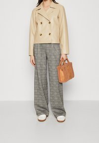 Vrouw in een beige dubbelzijdige jasje, grijs geruite wijde broek, witte sneakers, en met een kleine bruine fuzzy tote bag.