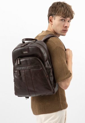 Mladý muž v hnědém pleteném tričku a krémových kalhotách, nesoucí tmavě hnědý kožený batoh na jednom rameni, dívá se stranou.