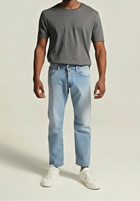 Homme portant un t-shirt gris à manches courtes, jean bleu clair et baskets blanches, debout sur un fond uni.