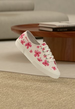 Hvit lerretssneaker med rosa blomsterprint og hvite lisser, plassert nær et rundt trebord med stablede bøker i en nøytral stue.