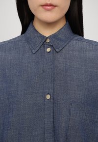 Femme portant une chemise chambray bleue avec un col pointu et des boutons-pression en métal, montrant l'encolure et le haut de la poitrine.