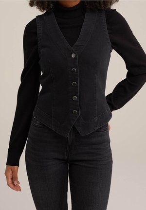 Frau trägt eine taillierte schwarze Weste mit Knöpfen über einem schwarzen Langarm-Rollkragenpullover und schwarzen Jeans vor neutralem Hintergrund.