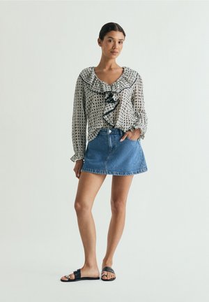 Jonge vrouw die een geplooide blouse met patroon, een denim minirok en zwarte slippers draagt, staand tegen een effen achtergrond.