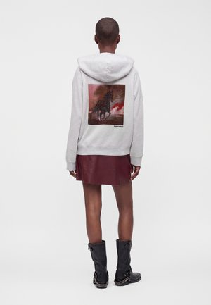 Personne portant un sweat à capuche gris avec un motif cheval noir dans le dos, associé à une jupe bordeaux et des bottes noires avec des détails en chaîne, vue de dos.