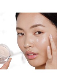 Clear glass jar containing a creamy, light beige moisturizer; model applying product to skin with a smooth, glossy finish. Neutral-toned background.