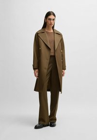 Cappotto trench doppiopetto marrone, con ampi revers, abbinato a un maglione marrone a coste e pantaloni coordinati, completato da scarpe nere.