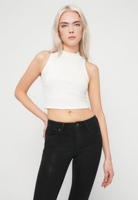 Fiatal nő hosszú szőke hajjal, fehér ujjatlan crop toppot és fekete, magas derekú farmert viselve, egy sima világos háttér előtt áll.