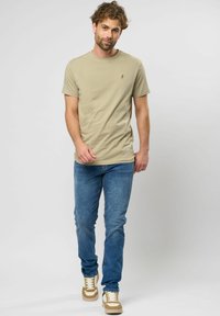 Beige pamut póló kerek nyakkal és egy kis logóval, kék farmerral és világosbarna sportcipővel párosítva, fehér fűzőkkel és csíkokkal.