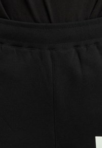 Schwarzer elastischer Bund an einer Jogginghose, die einen glatten Stofftextur und ein weißes Logodesign am unteren Bein aufweist.