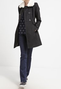 Manteau noir avec un col en fausse fourrure, conception à double boutonnage et deux poches latérales, associé à un pantalon sombre et des baskets texturées.