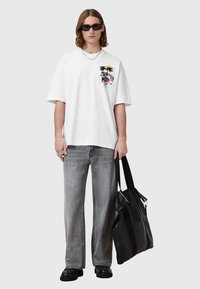 Hvit oversized t-skjorte med grafisk design, kombinert med grå vide jeans og svarte skinnsko. Svart totebag holdes i den ene hånden.