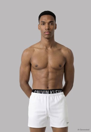 Giovane uomo con corporatura muscolosa che indossa pantaloncini bianchi Calvin Klein su sfondo grigio semplice, rivolto verso avanti con le braccia dietro la schiena.