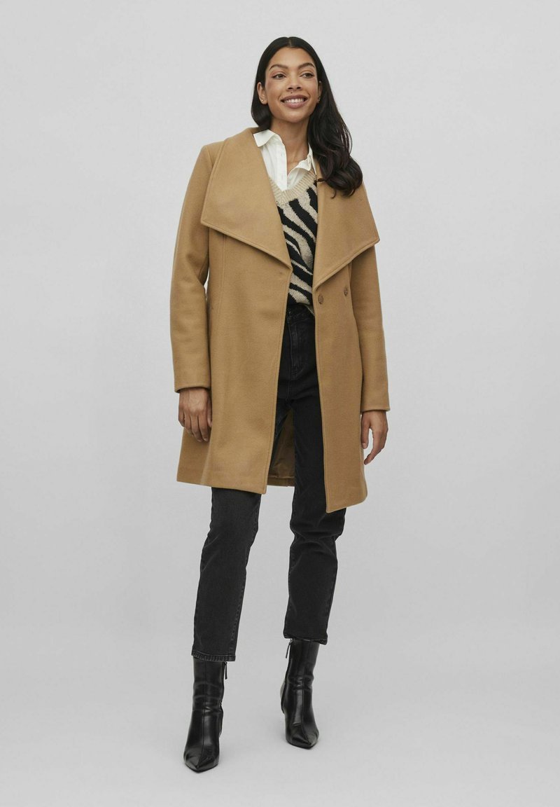 Abrigo de mezcla de lana color camel con solapa ancha, cierre de un solo botón, silueta oversize, llevado sobre un suéter de rayas negro y beis y unos jeans negros.