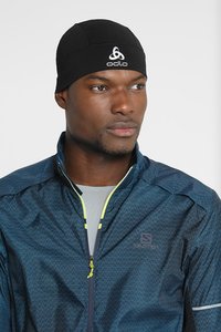 Zwarte sportieve beanie met Odlo-logo. Model draagt een marineblauwe jack met ritssluiting, voorzien van een geometrisch patroon en neon groene accenten.